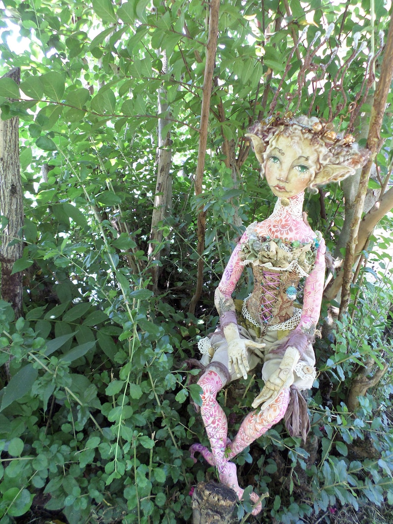 May include: A whimsical doll with a pink and white patterned dress, sitting in a forest setting. The doll has a crown of leaves and flowers on its head and is wearing a corset with lace and beads.