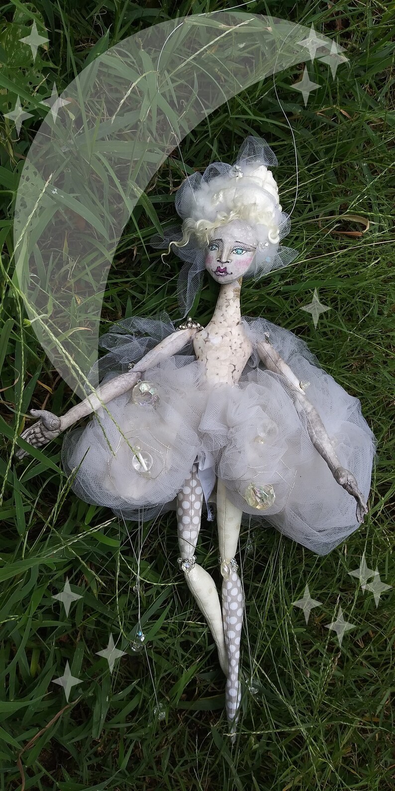 May include: A white fabric doll with a white tulle skirt and white polka dot tights. The doll is posed with its arms outstretched and is surrounded by green grass and a crescent moon.