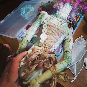 May include: A handmade doll with a green and white patterned body, pink hair, and a white dress with a brown ribbon and buttons. The doll is wearing a pair of white wings.