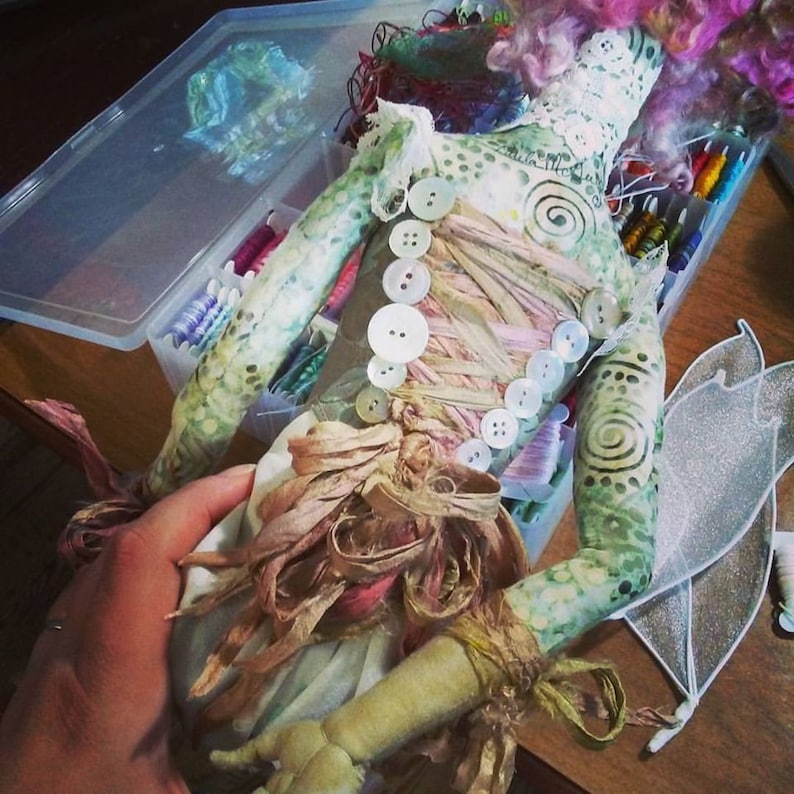 May include: A handmade doll with a green and white patterned body, pink hair, and a white dress with a brown ribbon and buttons. The doll is wearing a pair of white wings.