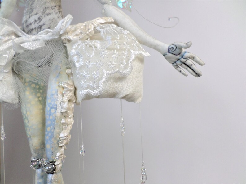 May include: A white porcelain doll with a white lace dress and a white fabric bag. The doll has a white painted face and a hand with a blue and black swirl design.