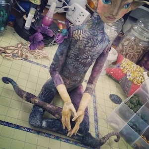 May include: A handmade doll with a purple and blue patterned body, long limbs, and a painted face. The doll is sitting with its legs crossed and its hands clasped together. The doll is on a table with a checkered surface.