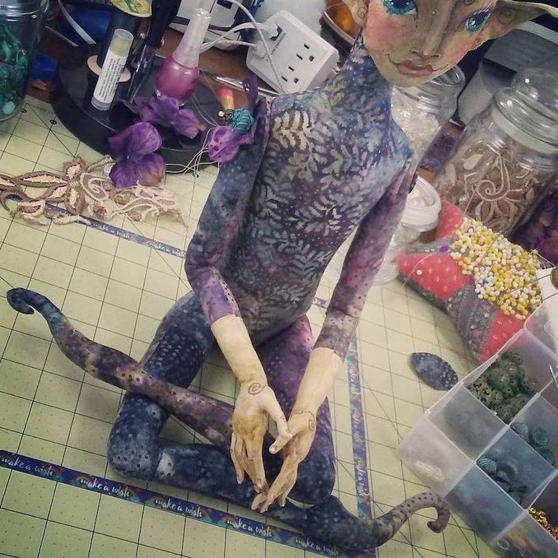May include: A handmade doll with a purple and blue patterned body, long limbs, and a painted face. The doll is sitting with its legs crossed and its hands clasped together. The doll is on a table with a checkered surface.