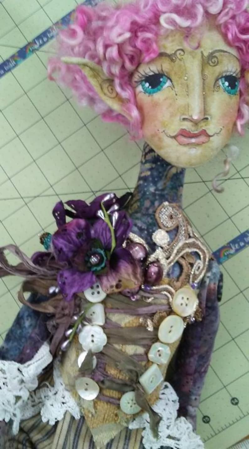 May include: A whimsical doll with pink curly hair, large blue eyes, and a painted face. The doll is wearing a purple flower, a brown and beige fabric bodice with pearl buttons, and a white lace skirt.