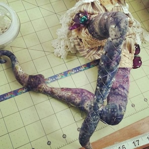 May include: A close-up of a doll's legs made of fabric and decorated with rhinestones. The legs are purple and black with a white and brown pattern. The doll is lying on a green and white checkered surface with a ruler in the foreground.