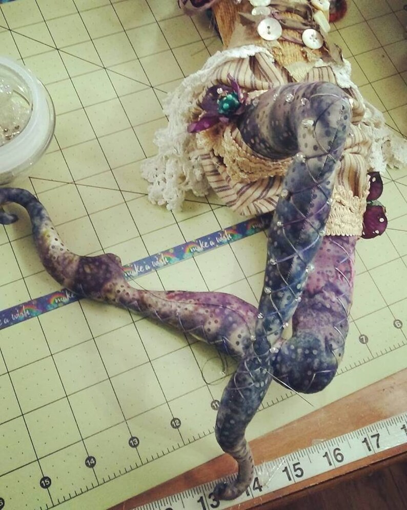May include: A close-up of a doll's legs made of fabric and decorated with rhinestones. The legs are purple and black with a white and brown pattern. The doll is lying on a green and white checkered surface with a ruler in the foreground.