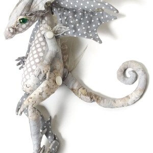 May include: A gray and white fabric dragon with green eyes and a curled tail. The dragon has white polka dots on its wings and body.