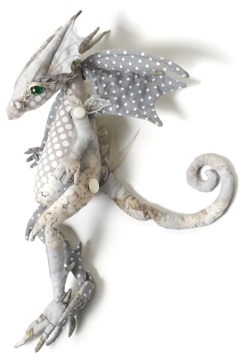 May include: A gray and white fabric dragon with green eyes and a curled tail. The dragon has white polka dots on its wings and body.