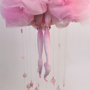 May include: A pink tulle ballerina mobile with crystal beads and pink glass beads hanging from the bottom. The ballerina is made of fabric and has pink legs and a pink tulle skirt.