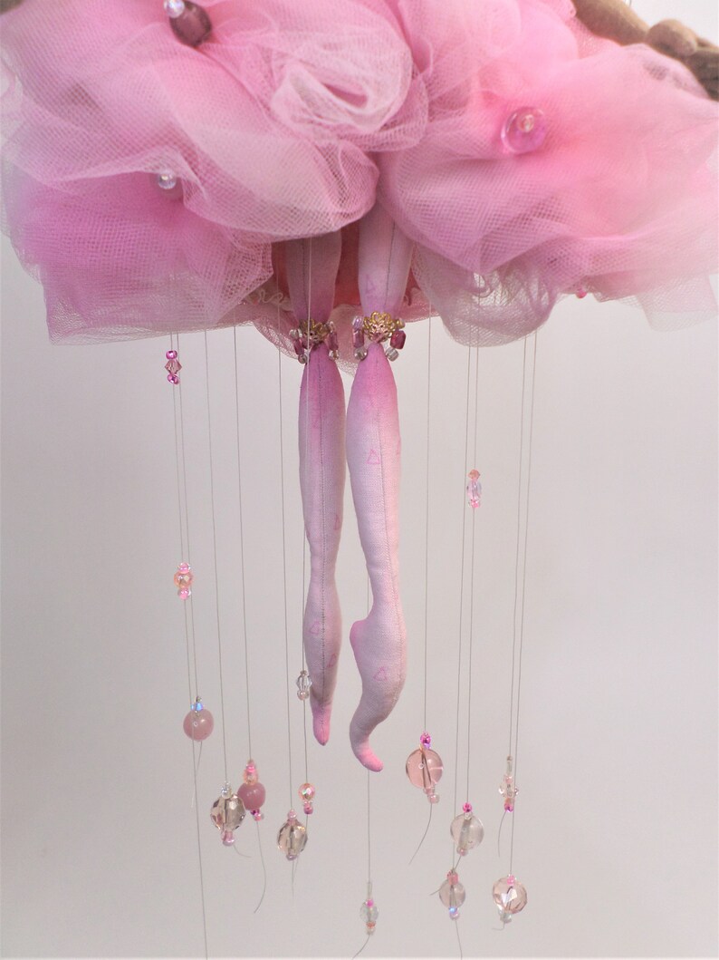 May include: A pink tulle ballerina mobile with crystal beads and pink glass beads hanging from the bottom. The ballerina is made of fabric and has pink legs and a pink tulle skirt.