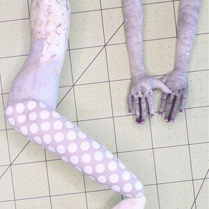 May include: A handmade doll with a gray and white patterned body, white polka dot legs, and gray arms. The doll is lying on a green cutting mat.