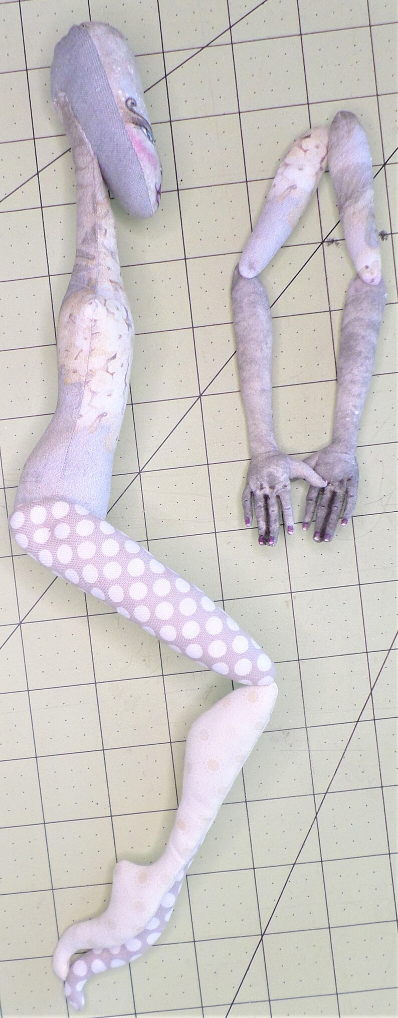 May include: A handmade doll with a gray and white patterned body, white polka dot legs, and gray arms. The doll is lying on a green cutting mat.