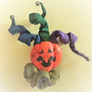 Puede incluir: Una calabaza naranja hecha a mano con una cara de calabaza tallada pintada, que lleva un sombrero de bufón verde, azul y morado con cascabeles.
