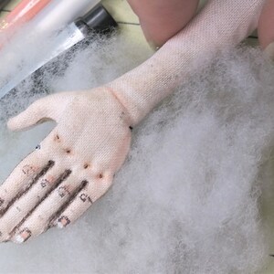 May include: A close-up of a white fabric hand with stitched details, including fingers and nails. The hand is resting on a bed of white fluffy stuffing.