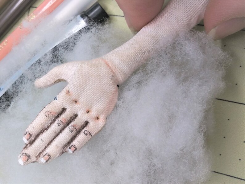 May include: A close-up of a white fabric hand with stitched details, including fingers and nails. The hand is resting on a bed of white fluffy stuffing.
