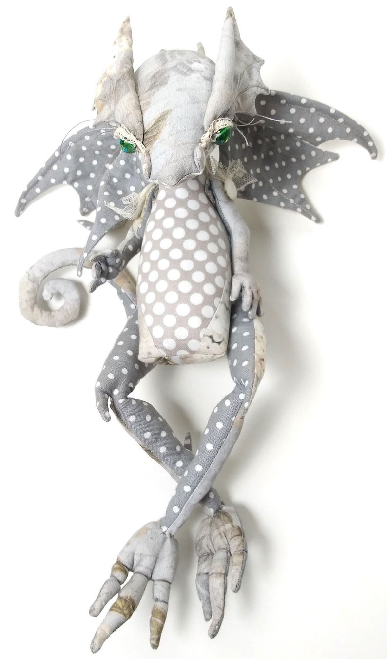May include: A gray and white fabric dragon with green eyes and polka dot wings. The dragon has a curled tail and long, thin legs.