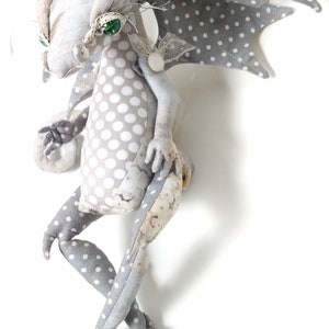 May include: A gray and white polka dot fabric dragon with green eyes and a white lace accent. The dragon has a long tail and four legs.