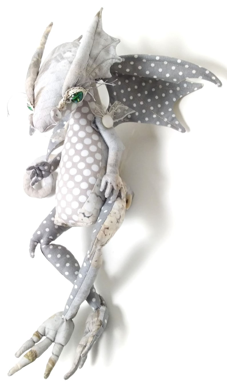 May include: A gray and white polka dot fabric dragon with green eyes and a white lace accent. The dragon has a long tail and four legs.