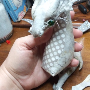 May include: A white and gray fabric dragon with a green eye and white polka dots. The dragon has large ears and a long tail.