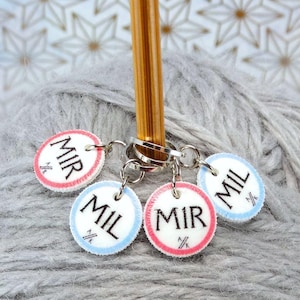 May include: Four round, white stitch markers with blue and red trim. The markers are labelled "MIL" and "MIR" and are attached to a wooden knitting needle.