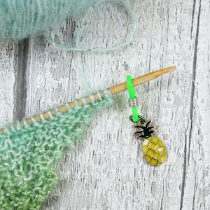 May include: A close-up of knitting needles with a green and yellow pineapple charm. The knitting project is a gradient of green and blue yarn with silver thread. The needles are made of wood.