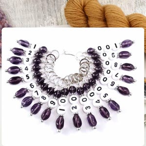 May include: A set of 10 purple and silver stitch markers with white numbered beads. The beads are numbered 0-9. The markers are connected by a silver ring.