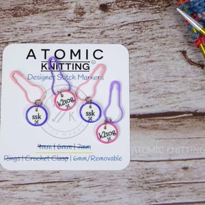 May include: A pack of Atomic Knitting designer stitch markers. The markers are in shades of pink and purple, with the text "ssk" and "k2tog" on the circular charms. The package also includes the text "Rings | Crochet Clasp | 6mm/Removable".