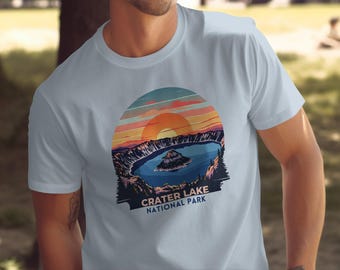 May include: A light blue t-shirt with a vintage-style graphic of Crater Lake National Park. The graphic features a sunset over the lake with a small island in the center.