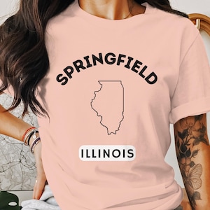 Puede incluir: Una camiseta rosa con un contorno negro del estado de Illinois y el texto "SPRINGFIELD ILLINOIS" impreso en la parte delantera.