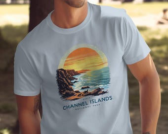 Op de afbeelding: Een lichtblauw t-shirt met een afbeelding van een zonsondergang boven een rotsachtige kustlijn en de tekst "Bezoek Channel Islands National Park".