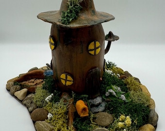 May include: A miniature fairy house crafted from brown clay, featuring a pointed roof and a mushroom-shaped chimney. The house is nestled amongst moss, small stones, and white flowers.