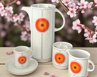 May include: A white ceramic coffee set with a retro design. The set includes a tall coffee pot, a small cup and saucer, a creamer, and a sugar bowl. Each piece features a large orange and yellow circular design with black accents.