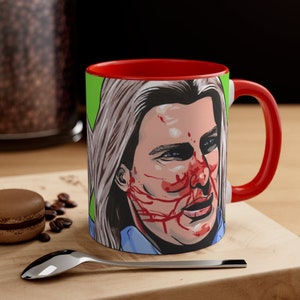 May include: A red and white coffee mug with a graphic of a person's face with blood splatters on it. The person has long blonde hair and is wearing a blue shirt.