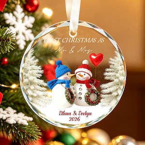 May include: A round, clear glass Christmas ornament featuring two snowmen, one with a blue hat and scarf and one with a red hat and scarf, holding small wreaths. The text reads FIRST CHRISTMAS AS Mr & Mrs, Ethan & Evelyn, and 2026.