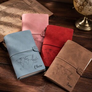 May include: Four rectangular journals with matte covers in light blue, pink, red, and brown. Each journal features a world map outline, a small airplane icon, a dotted travel path, and a name printed in script: Olivia and Luna.