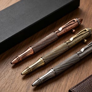May include: Three metal bolt-action pens in copper, brass, and gunmetal finishes. Each pen features a textured, twisted barrel design, a pocket clip, and a side bolt mechanism. The brass pen is engraved with the text THIS IS FOR YOU.