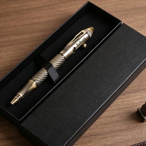 May include: A metallic bronze-toned ballpoint pen with a spiraled grip and a clip, featuring the engraved name Sylvan on the barrel. The pen is secured by a black strap inside a rectangular black gift box, accompanied by a small bronze-toned seal.