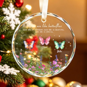Könnte beinhalten: Ein rundes, klares Glasornament, das an einem weißen Band hängt. Es zeigt den Text "Sisters are like butterflies. EACH ONE IS DIFFERENT. EACH ONE IS BEAUTIFUL. EACH ONE IS SPECIAL." mit drei Schmetterlingen namens "Matilda," "Clara," und "Stella," über einem bunten Feld aus Wildblumen.