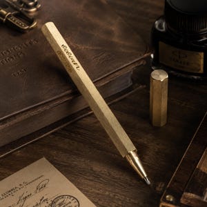 May include: A hexagonal brass pen featuring the engraved name "Lilian" and the date "08.22.2026" on its side. The pen has a metallic finish, a pointed tip, and a separate matching hexagonal cap resting nearby, showcasing a clean and functional design.