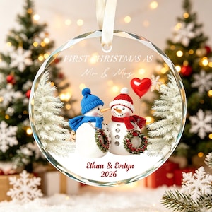May include: A round, clear glass Christmas ornament featuring a printed graphic of two snowmen. One snowman wears a blue hat and scarf, the other a red hat and scarf. The text reads FIRST CHRISTMAS AS Mr & Mrs, Ethan & Evelyn, and 2026.