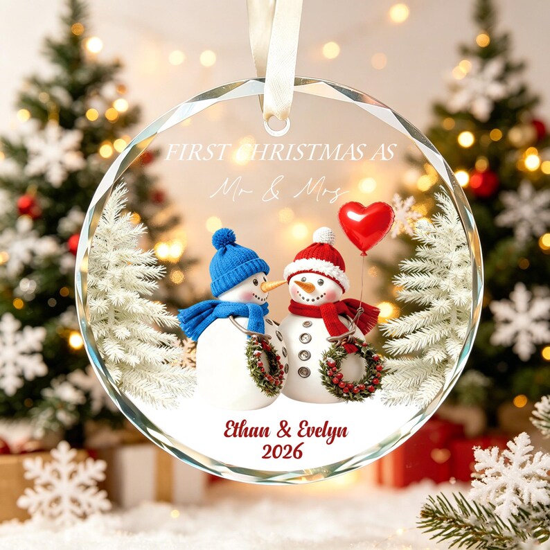 May include: A round, clear glass Christmas ornament featuring a printed graphic of two snowmen. One snowman wears a blue hat and scarf, the other a red hat and scarf. The text reads FIRST CHRISTMAS AS Mr & Mrs, Ethan & Evelyn, and 2026.