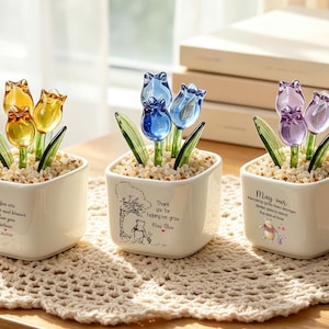 Puede incluir: Tres macetas cuadradas de cerámica blanca con tulipanes de cristal en amarillo, azul y morado. Cada maceta tiene hojas de cristal verde, pequeñas piedras y texto impreso con ilustraciones de un oso. El texto incluye citas y fechas.