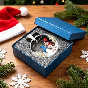 May include: A round glass Christmas ornament featuring two snowmen, one with a red scarf and one with a blue scarf, holding a red heart. The text reads First Christmas as Mr & Mrs and Eilun & Edlyn 2026. It is presented in a blue gift box.