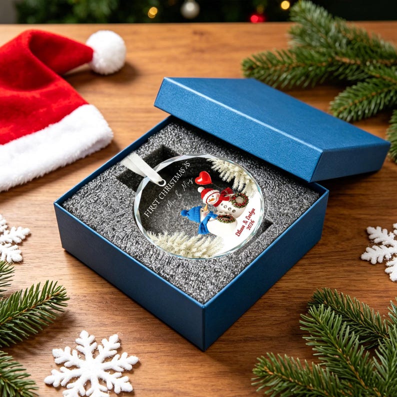 May include: A round glass Christmas ornament featuring two snowmen, one with a red scarf and one with a blue scarf, holding a red heart. The text reads First Christmas as Mr & Mrs and Eilun & Edlyn 2026. It is presented in a blue gift box.