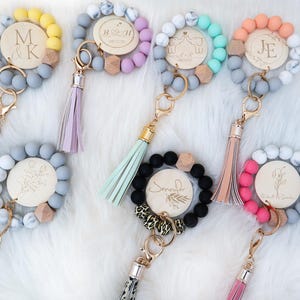 May include: A collection of personalized keychains with wooden engraved accents. Each keychain features a beaded bracelet design in various colors, including yellow, gray, pink, and black, with a tassel and gold-tone hardware. The wooden accents include initials and floral designs.