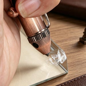 May include: A copper-toned metal tool with a textured, brushed finish and a pointed tip, positioned against the corner of a clear glass pane. The tool features a ridged grip section and a small metal clip, with a separate small metal component nearby.