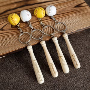 May include: Four baseball-themed keychains. Each keychain features a miniature baseball bat with a personalized name and a baseball. The baseballs are yellow and white, and the bats are light wood.