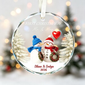 May include: A round, clear glass ornament featuring two snowmen, one with a blue hat and scarf and one with a red hat and scarf, holding a red heart balloon and wreaths. Text reads: FIRST CHRISTMAS AS Mr & Mrs, Ethan & Evelyn, 2026.