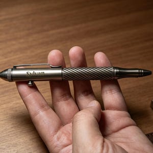 May include: A metallic bolt-action pen with a gunmetal finish, featuring a spiral-textured grip section, a pocket clip, and a pointed end cap. The barrel is engraved with the word Sylvan and includes a small side-mounted bolt mechanism.