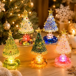 May include: Five miniature glass Christmas tree figurines with textured, crystalline surfaces and yellow star toppers. Each tree sits on a rounded, translucent base that glows with internal LED light in various colors including green, red, blue, and pink.